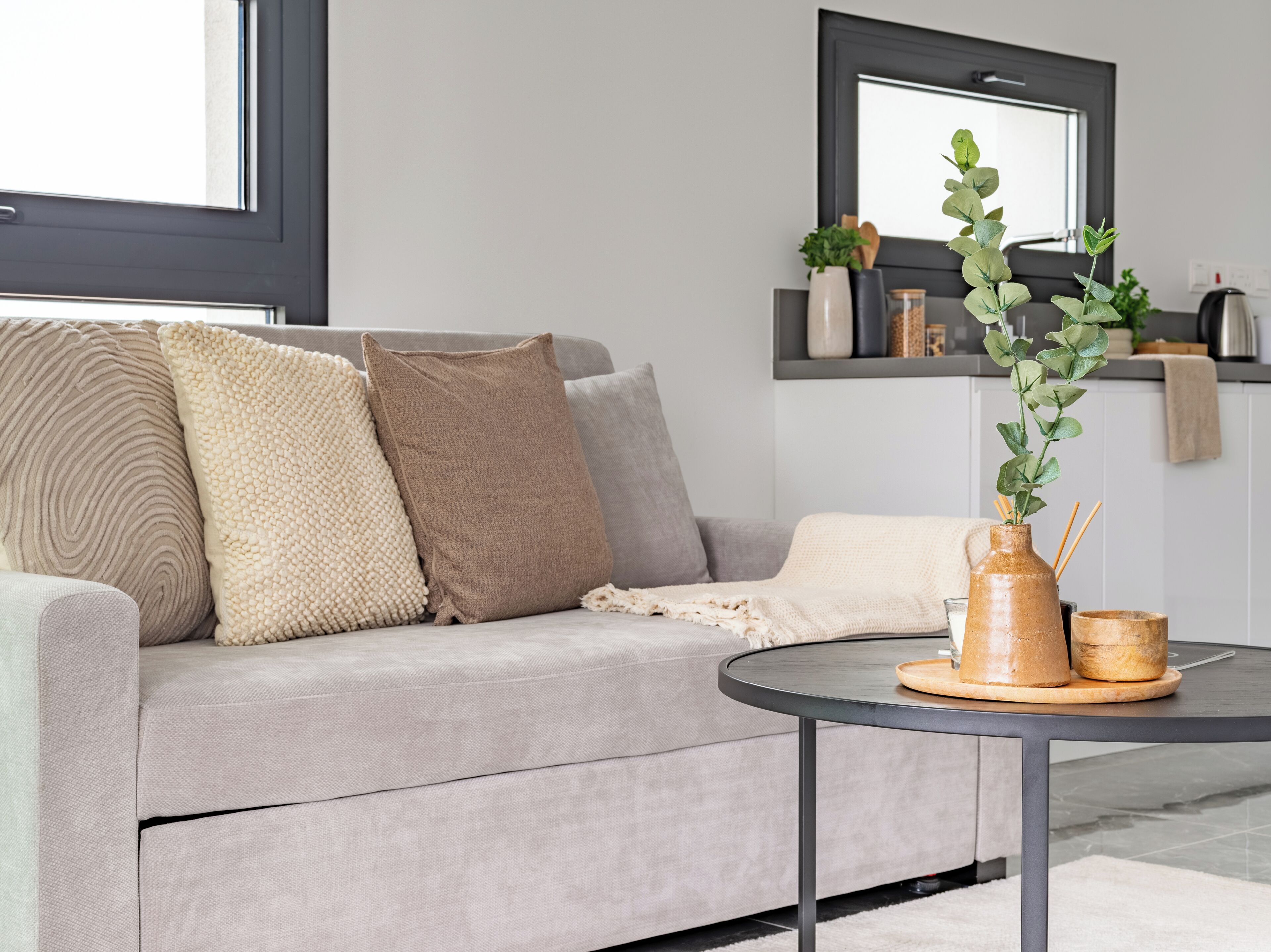 A modern living room with a grey sofa, multiple cushions, a round coffee table with a vase and candles, and a kitchen area in the background.