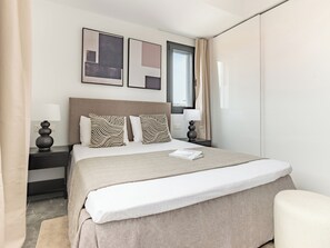 A modern hotel room with a large bed, two bedside tables with lamps, a window with curtains, and two framed artworks on the wall.
