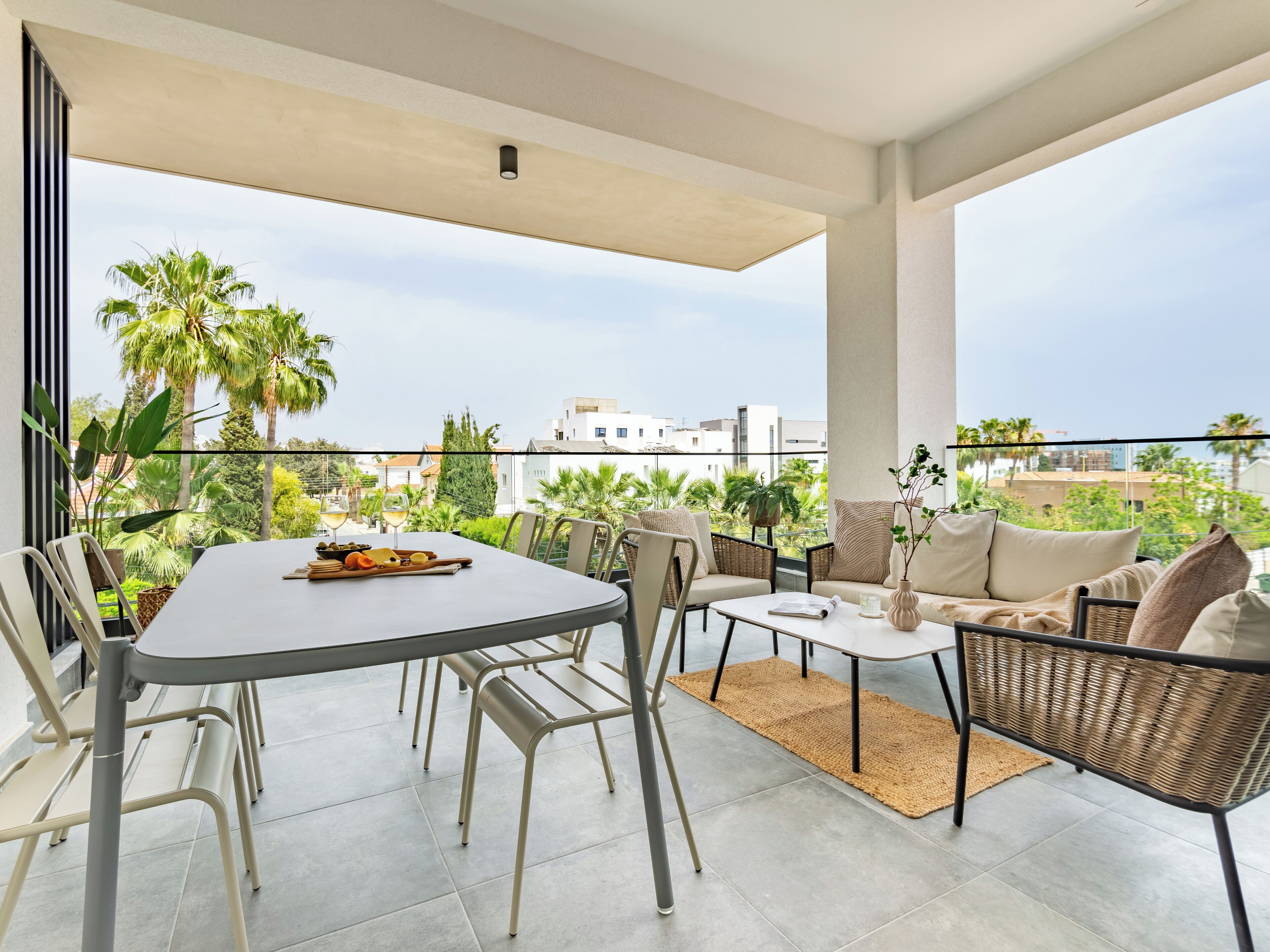 A spacious balcony with a dining table, chairs, and a sofa, overlooking a lush garden and cityscape.