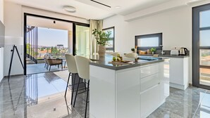 A modern kitchen with a large island, a dining area with chairs, and a view of the city through large windows. - Sanders Evolution 2 - 2 Bedroom Apartment With Private Rooftop (Limassol)