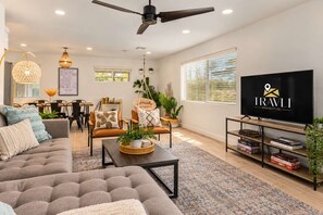 A modern living room with a sofa, coffee table, and TV. There is a dining area with chairs and a table. The room has large windows with blinds. - Vacay on Villa | Old Town |Swim, Putt, Play & Stay (Scottsdale)