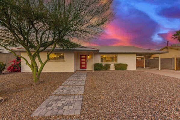 A single-story house with a red door, a gravel driveway, and a tree in the foreground. - Vacay on Villa | Old Town |Swim, Putt, Play & Stay (Scottsdale)