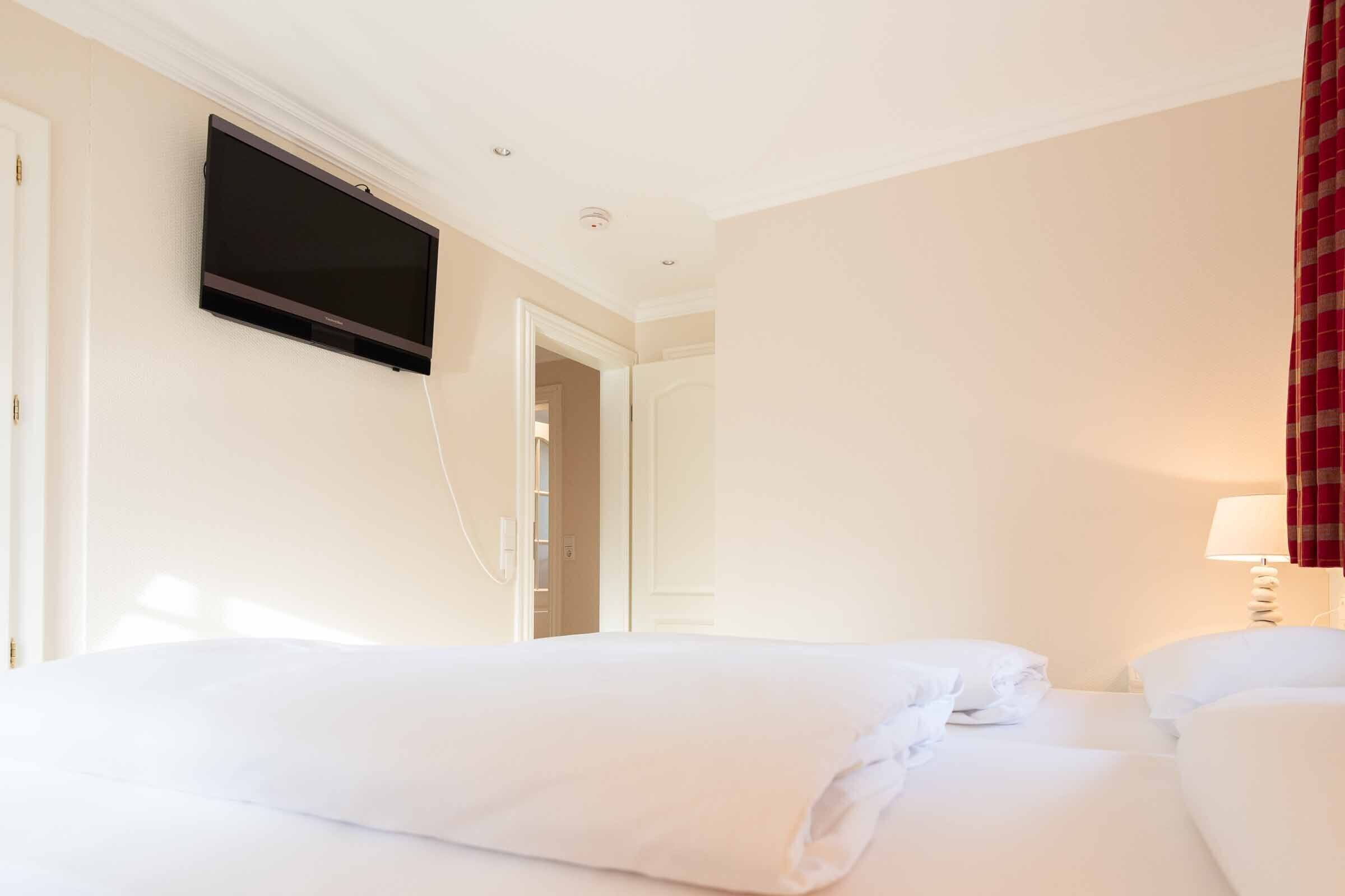 Et hotelværelse med et vægmonteret fladskærms-tv, en seng med hvide sengetøj og et natbord med en lampe.