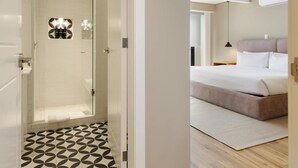 Ein modernes Hotelzimmer mit Holzboden, einem Bett mit Kopfteil sowie einem Badezimmer mit Dusche und einem gemusterten Boden.