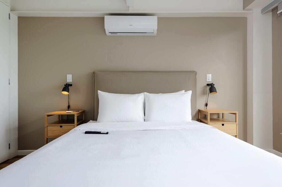 Una cama bien hecha con ropa blanca, dos mesitas de noche con lámparas y un aire acondicionado de pared.