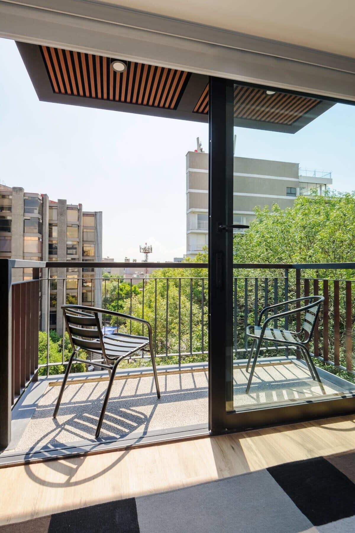 Un balcon avec deux chaises et une table, offrant une vue sur un paysage urbain composé d’immeubles modernes et d’espaces verts.