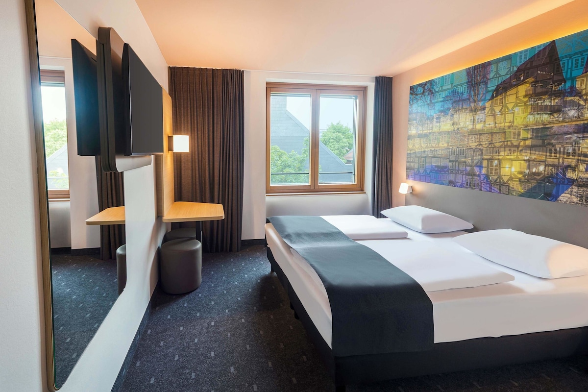 Una habitación de hotel con una cama, un escritorio, una silla, un cubo de basura, una ventana y un cuadro en la pared.