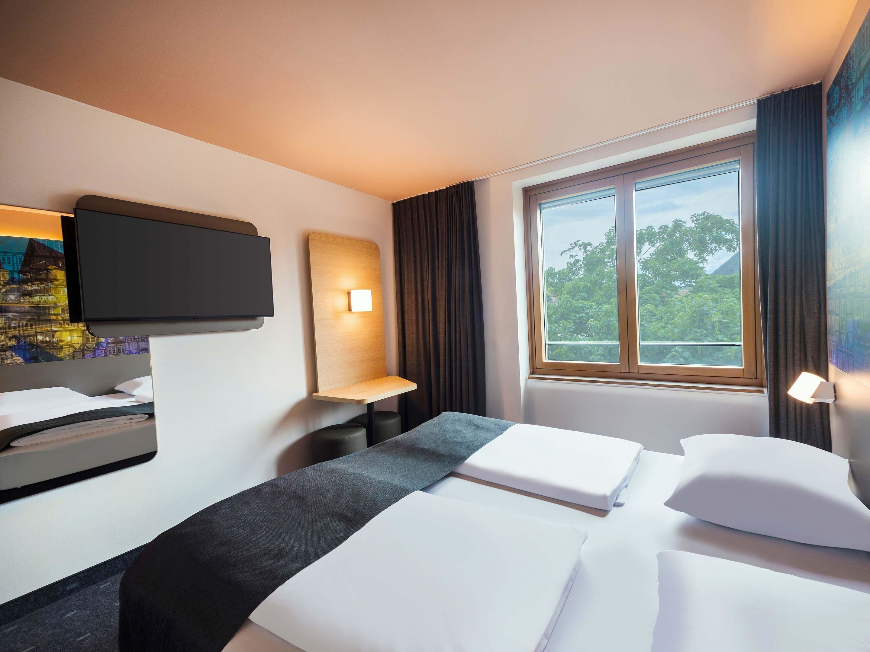 Ein Hotelzimmer mit Bett, Schreibtisch, einem an der Wand befestigten Flachbildfernseher und einem Fenster mit Blick auf Bäume.