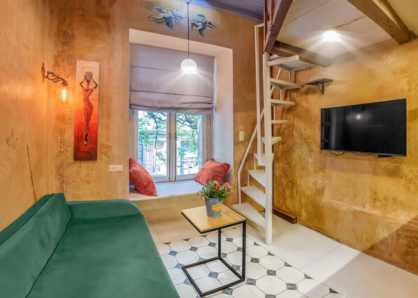 A living room with a green sofa, a small table with a vase, a staircase, and a TV mounted on the wall.