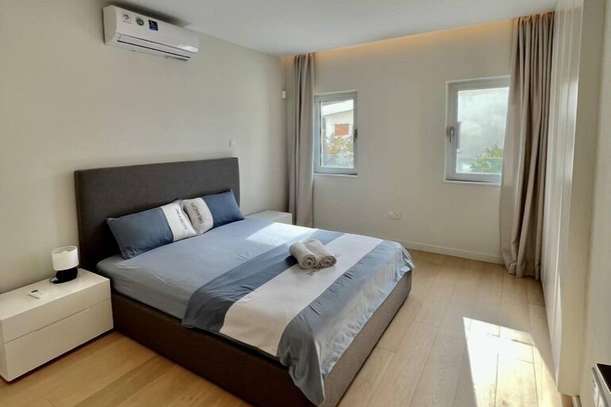 A bedroom with a bed, nightstand, air conditioning unit, and windows.