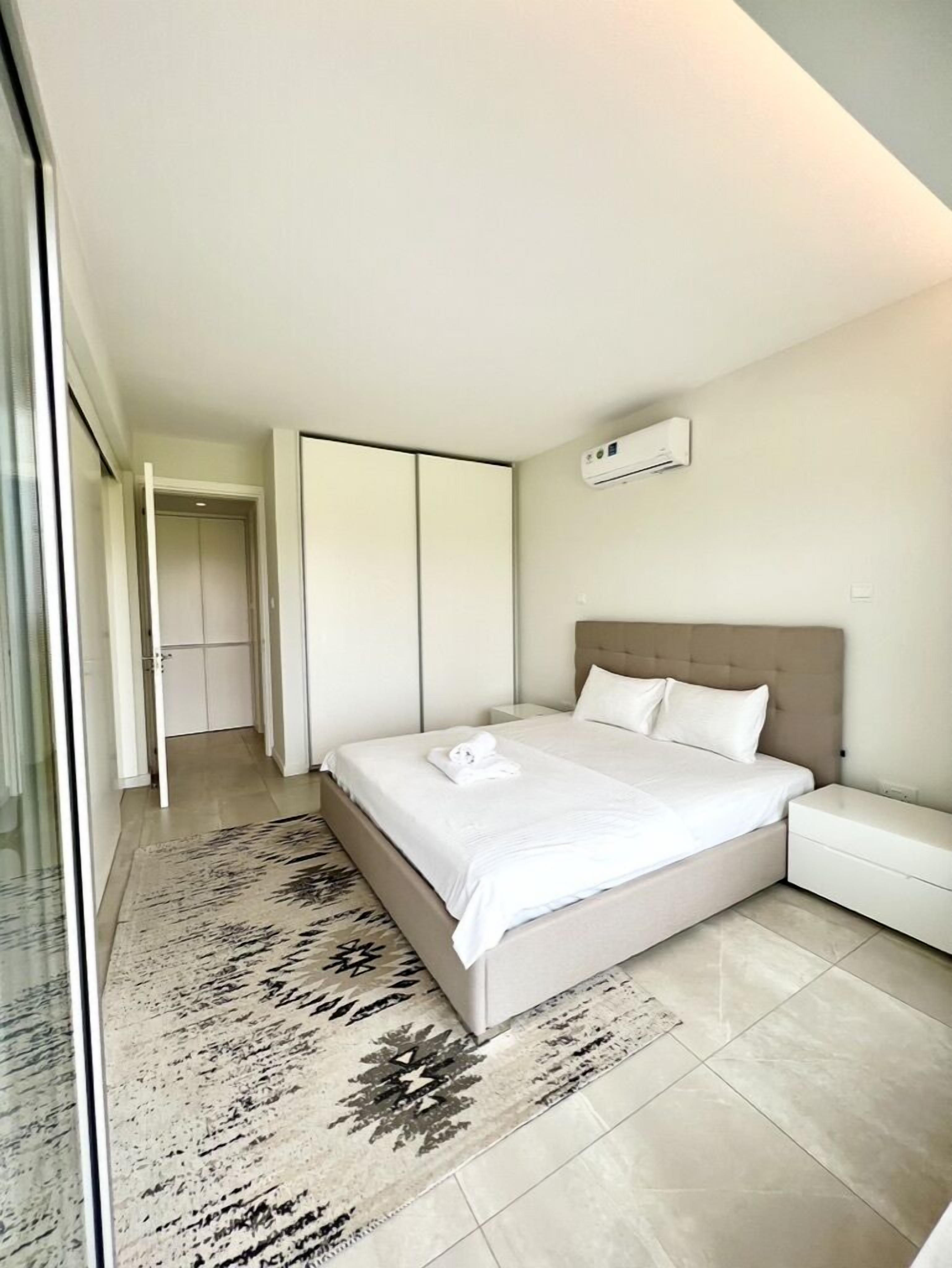 A modern bedroom with a large bed, a bedside table, a wall-mounted air conditioner, and a patterned rug.