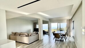 A spacious living area with a modern sofa, a dining table with chairs, and a large window offering a view. - Emerald Bay Suites (Paralimni)