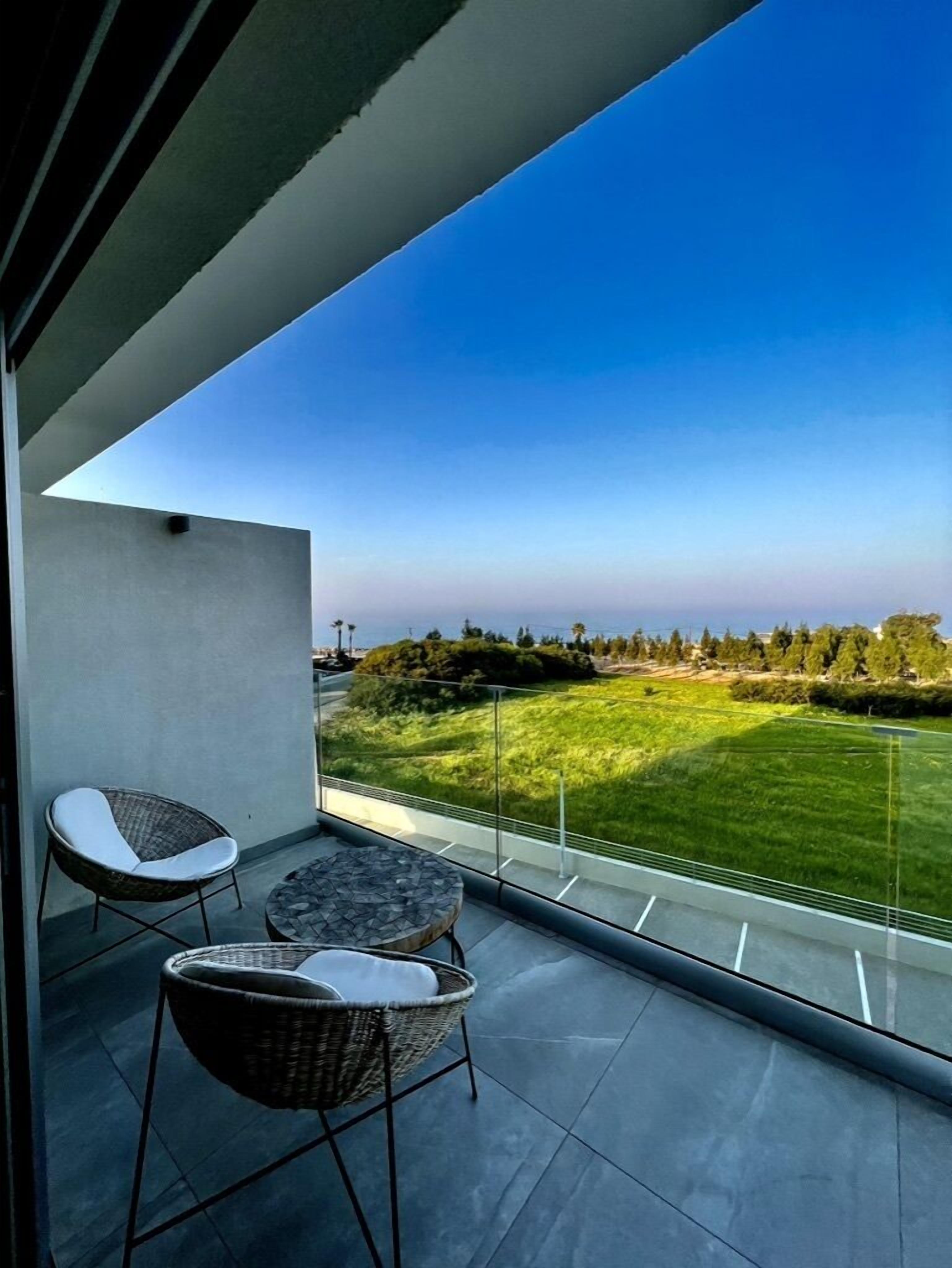A balcony with two wicker chairs and a small table, overlooking a grassy area with trees and a clear blue sky.