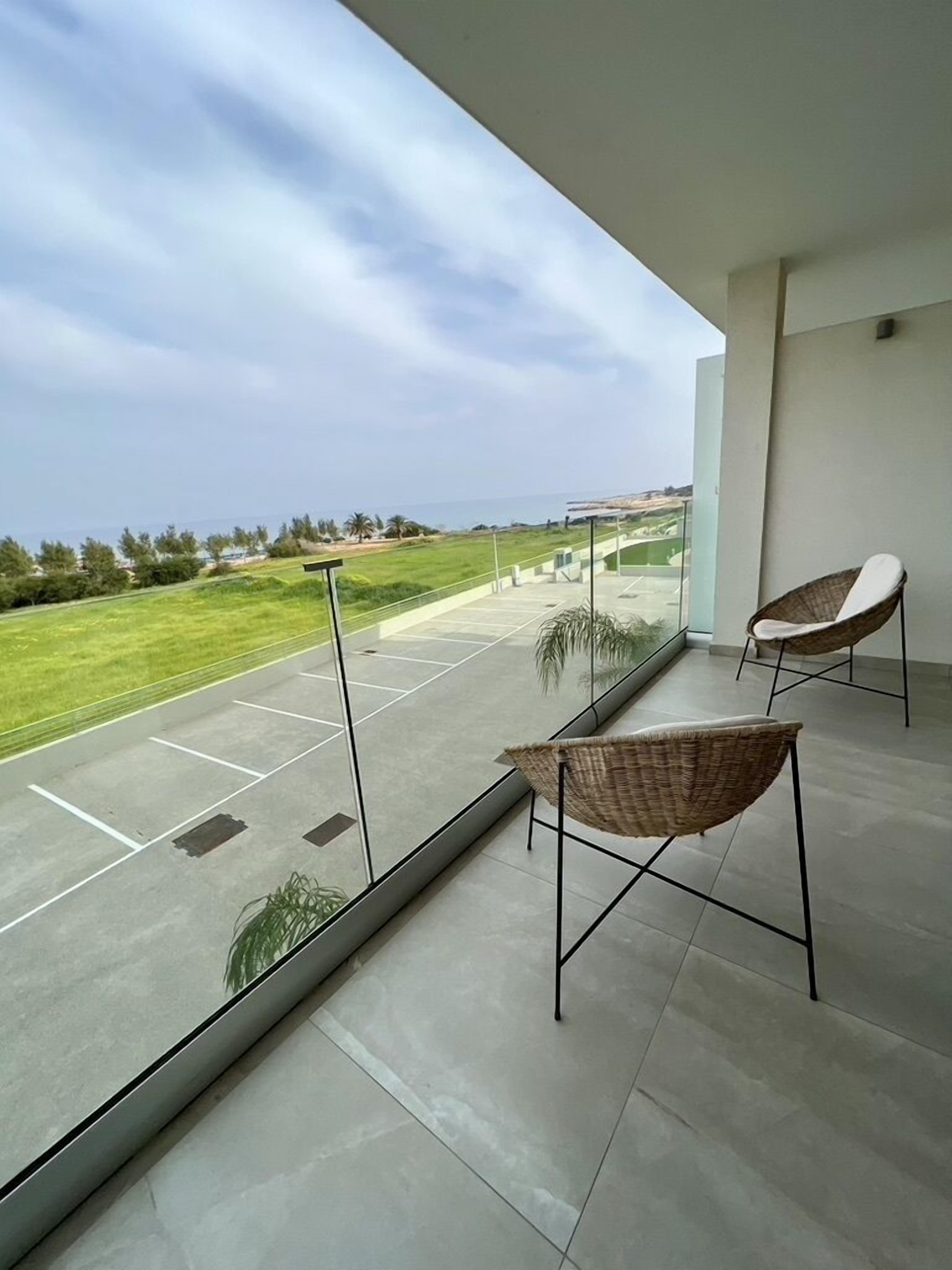 Un balcón con dos sillones de mimbre, barandilla de vidrio y vistas a un paisaje verde y colinas al fondo.