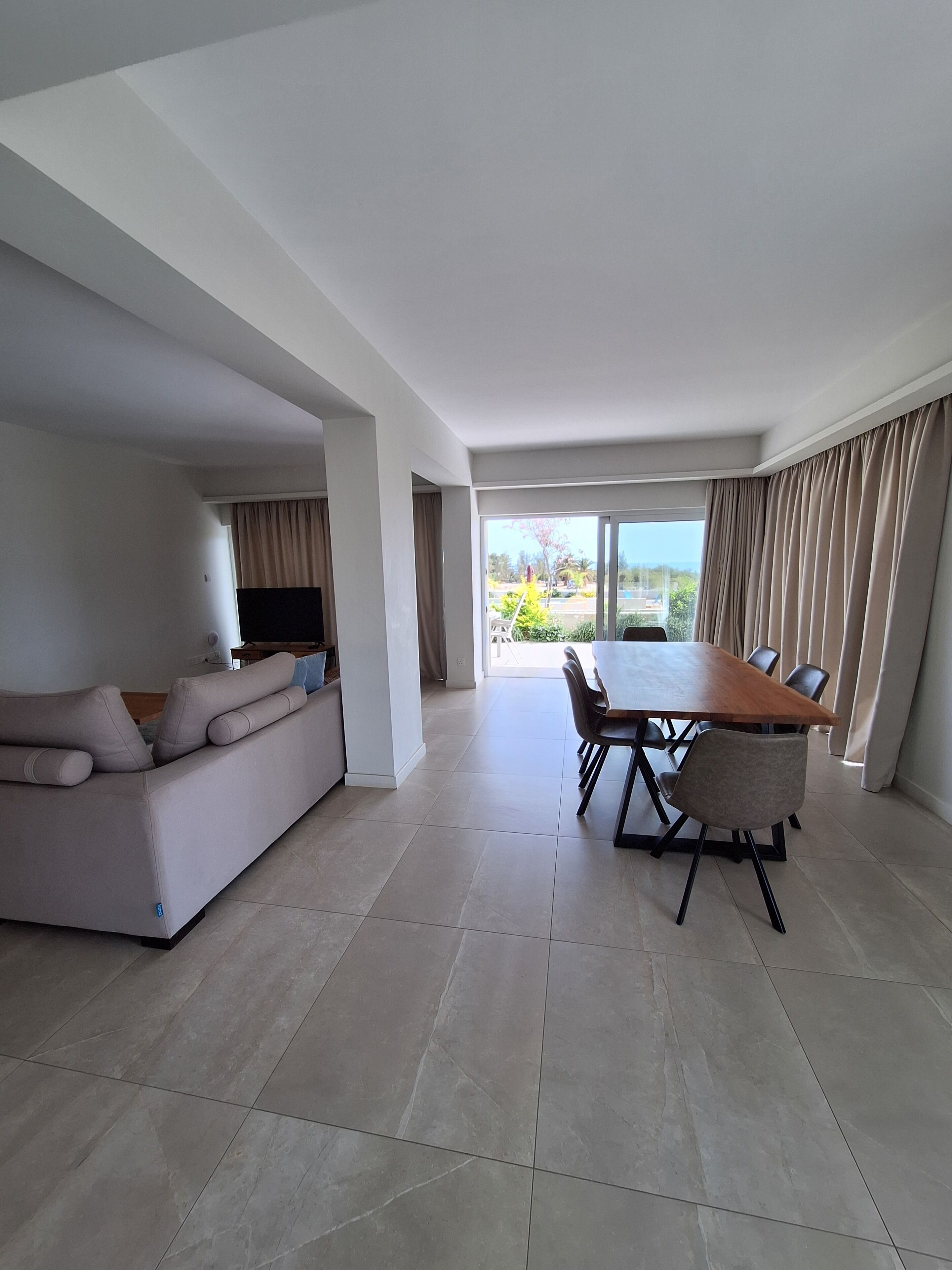 Uma sala de estar ampla com sofá, mesa de jantar e cadeiras, janelas grandes com cortinas e uma televisão de tela plana.