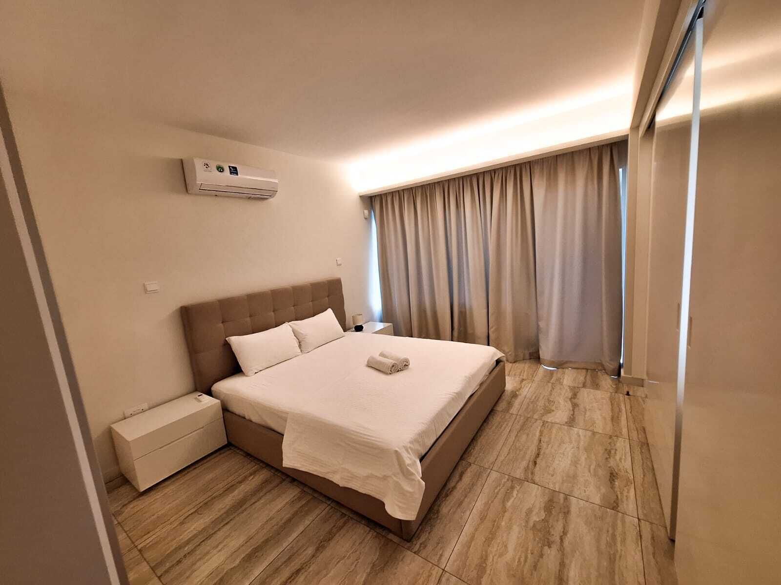 A hotel room with a bed, a nightstand, a bedside table, an air conditioning unit, and a window with curtains.