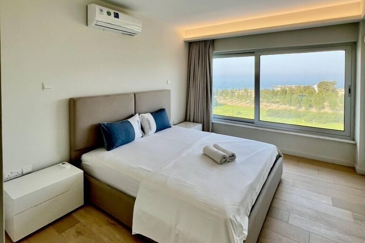 A hotel room with a bed, a nightstand, an air conditioning unit, a window with a view, and a ceiling light.