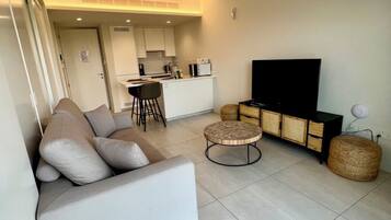 A modern living room with a sofa, a round coffee table, a flat-screen TV, and a wicker ottoman.