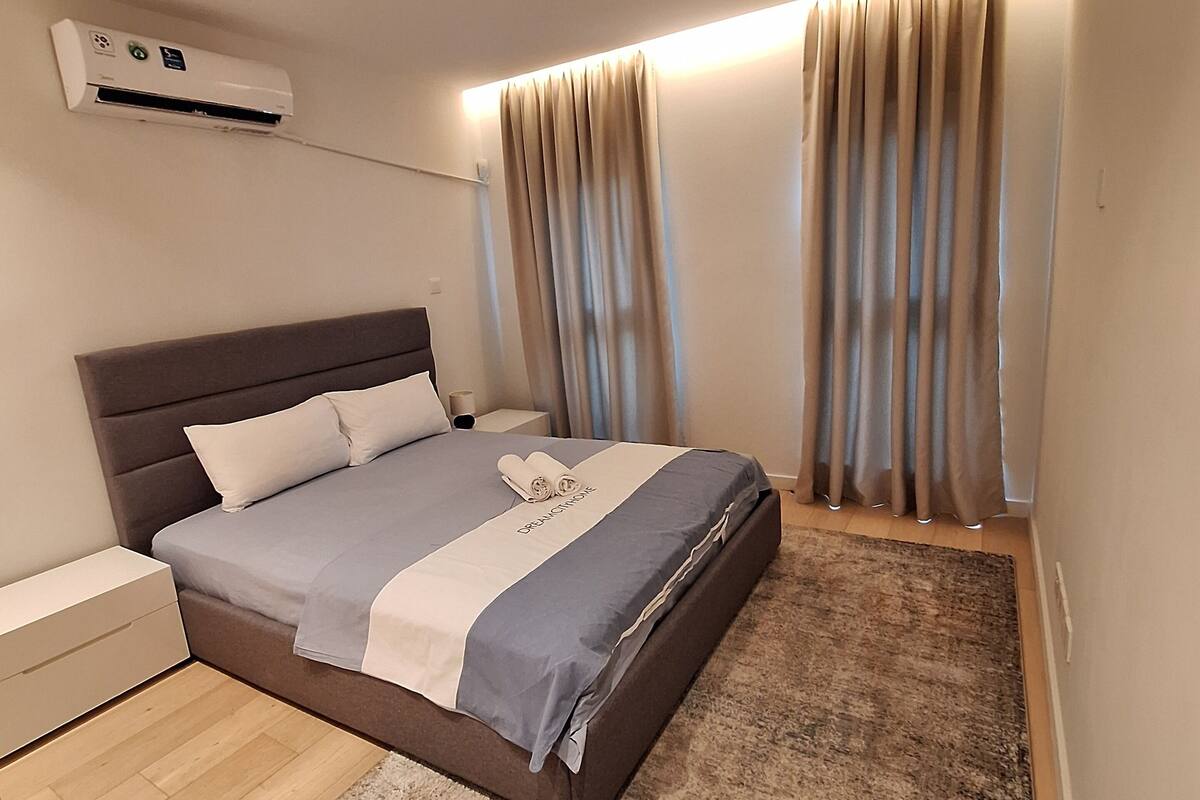 A modern bedroom with a bed, bedside table, air conditioning unit, and curtains.