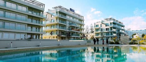 A modern hotel with multiple balconies, a swimming pool, and a clear blue sky.