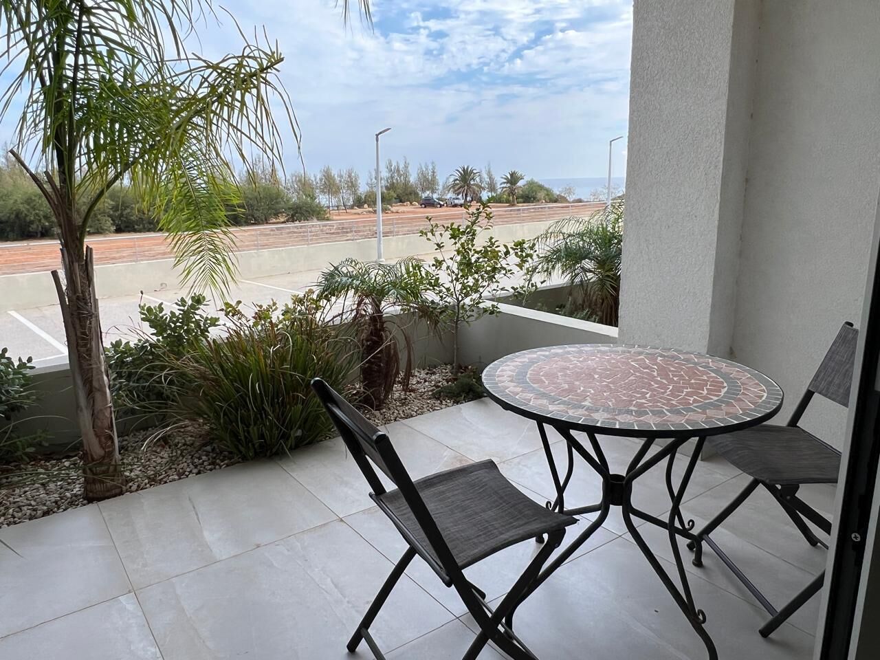 A patio with a round table and two chairs, a palm tree, and a view of a parking lot and distant landscape.