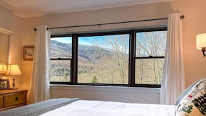 A bedroom with a large window offering a mountain view, a bed with a patterned pillow, and a bedside table with a lamp.
