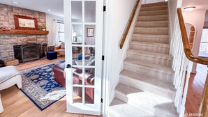 A spacious living room with a stone fireplace, a large open door leading to a staircase, and a cozy seating area with a blue rug.