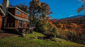 A wooden cabin with a deck and a view of autumn foliage and mountains.