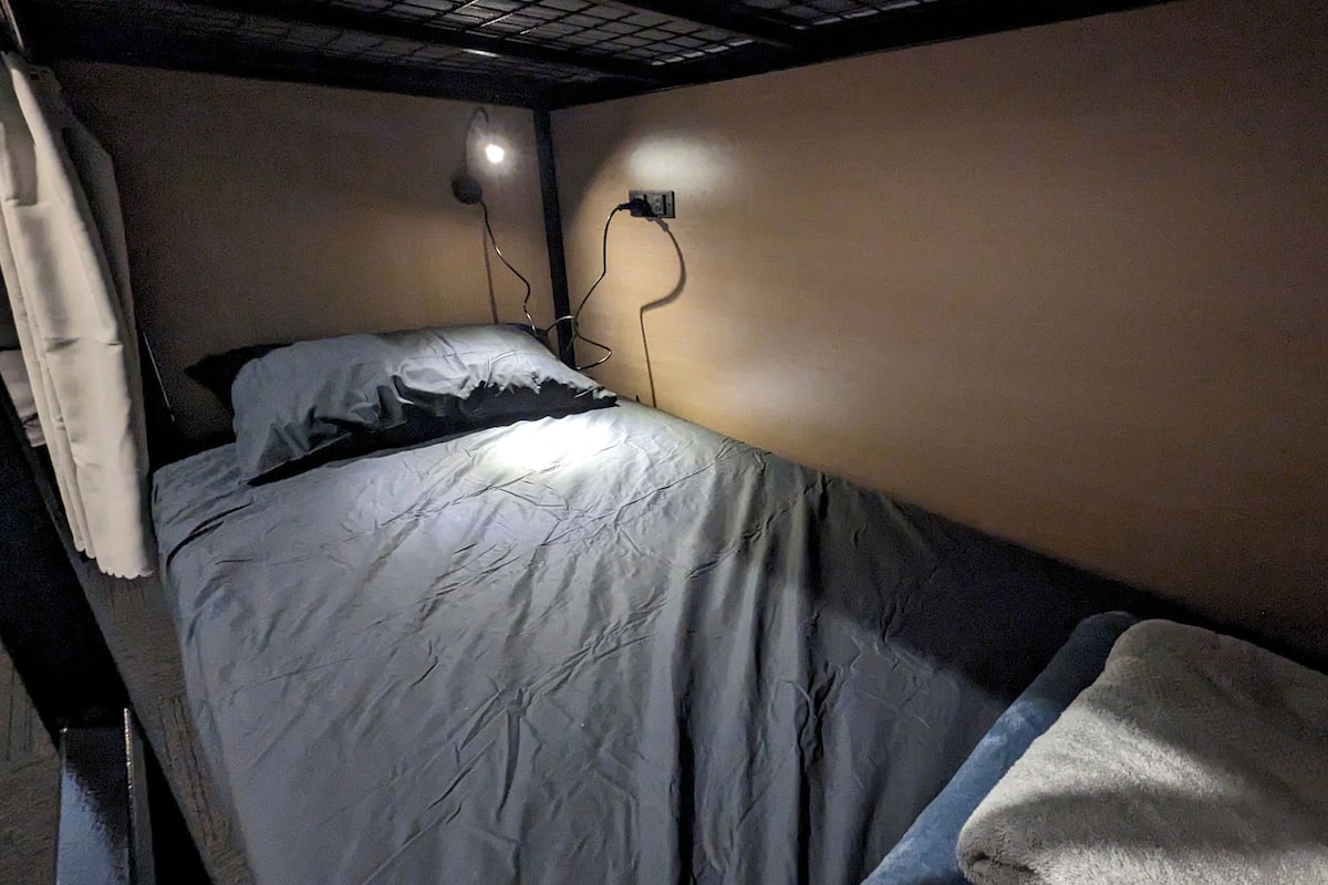 A single bed with a white blanket and a headboard, a wall socket, and a ceiling light fixture.