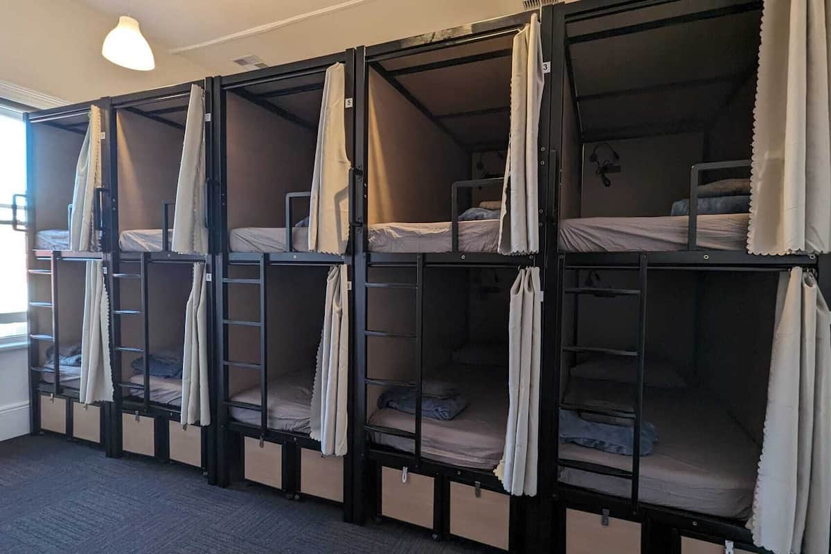 A dormitory room with bunk beds, each with a curtain, a small ladder, and a window.
