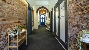 A corridor with brick walls, a wooden bench with plants, a door with a sign, and a wall-mounted light fixture.