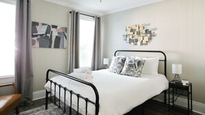 A neatly arranged bedroom with a bed, a bedside table, a chair, a ceiling fan, and a wall art piece.