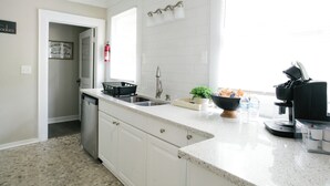 A kitchen with white cabinets, a dishwasher, a sink, a coffee maker, and a window with blinds.