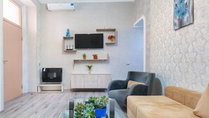 A modern living room with a glass coffee table, a beige sofa, a TV, and a wall-mounted shelf with decorative items. - H&B apartments Qolga (Tbilisi)