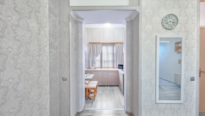 A narrow hallway with patterned wallpaper, a wooden bench, and a kitchen area with a window and curtains. - H&B apartments Qolga (Tbilisi)