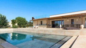Ein moderner Poolbereich im Freien mit klarem Pool, überdachter Terrasse mit Sitzgelegenheiten und einem Steinhaus im Hintergrund.