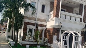A two-story building with a brick facade and white trim. There are potted plants on the ground floor and a white gate on the right side. - TAI HOTEL LEKKI (Lekki)