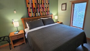 Ein Schlafzimmer mit einem großen Bett, einem Holzkopfschild, einem Nachttisch mit Lampe, einer Wandtäfelung und einem Fenster mit Blick auf Bäume.