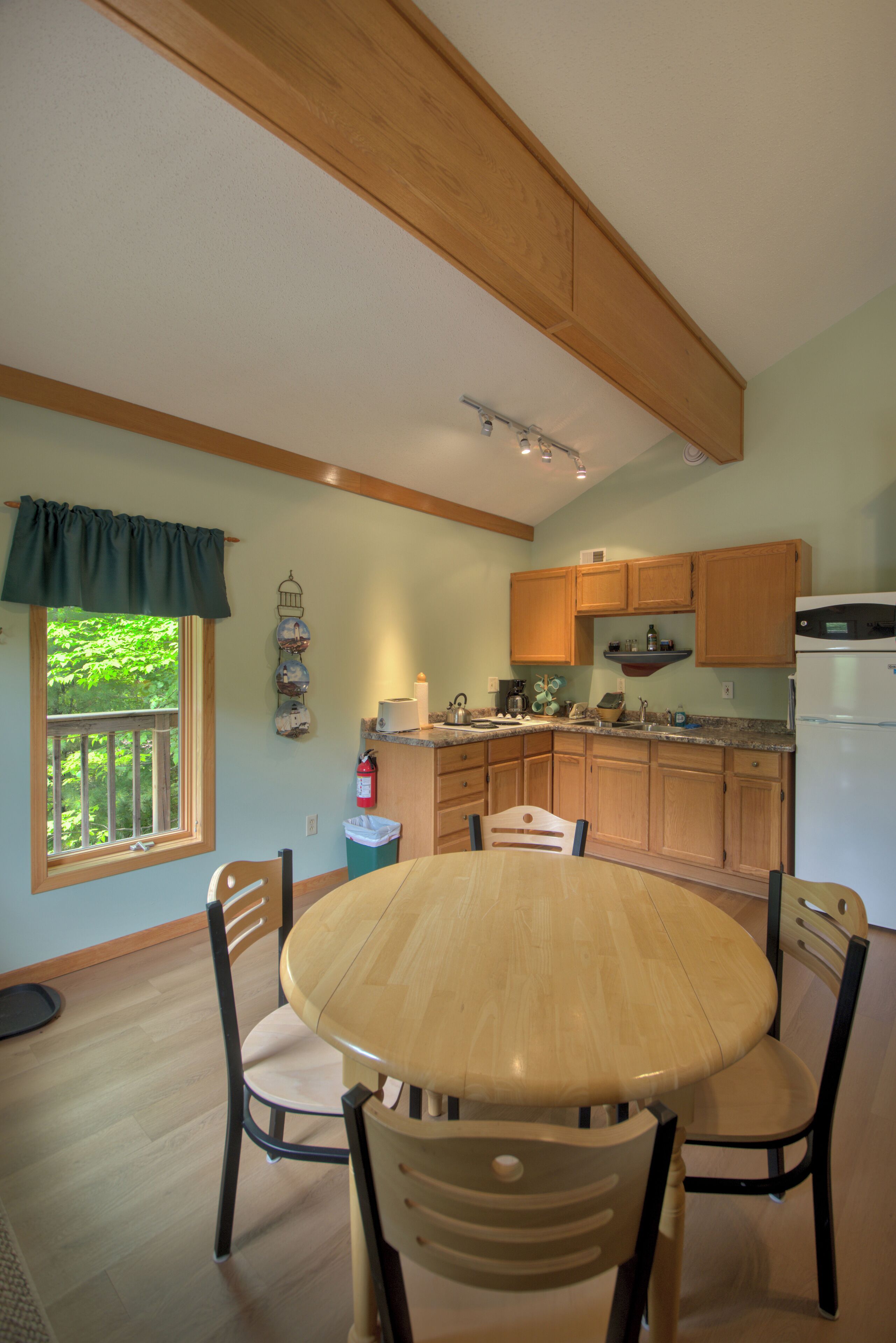 A kitchen with wooden cabinets, a dining table with chairs, a refrigerator, and a window with a view of greenery.