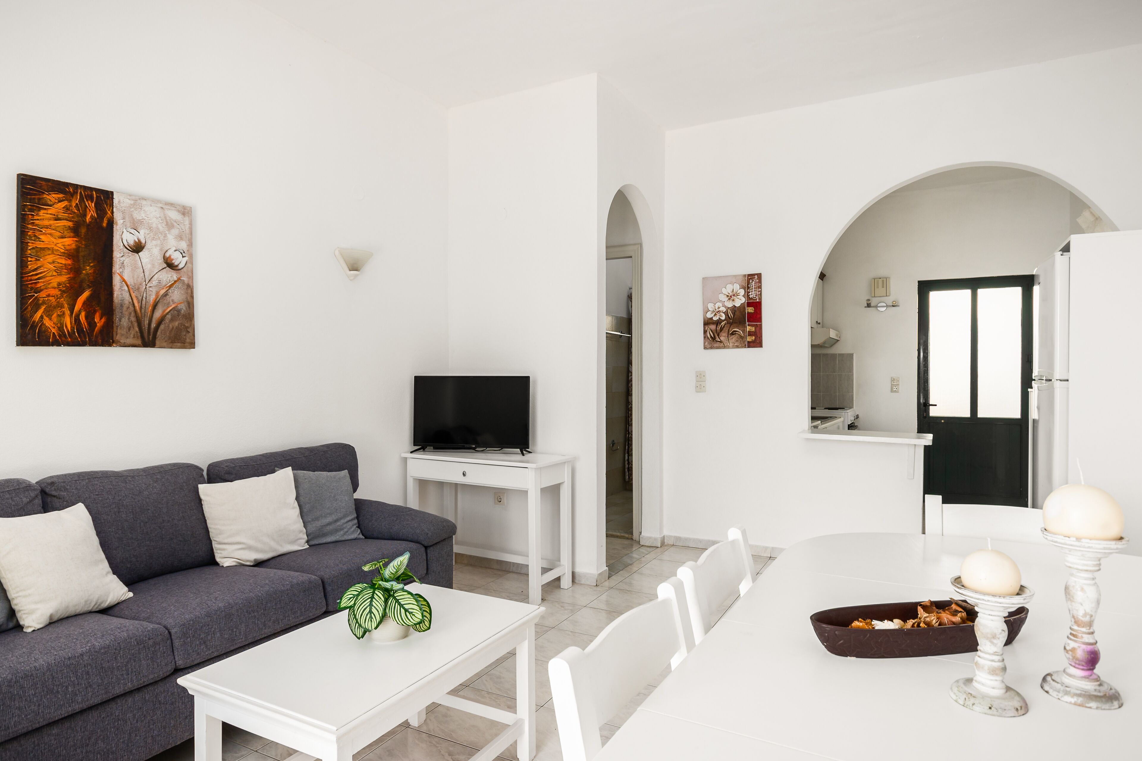 Un salon moderne avec un canapé gris, une table basse blanche et un téléviseur à écran plat posé sur un meuble blanc. On y trouve également un coin repas avec une table et des chaises blanches, et une cuisine visible à travers une arche.