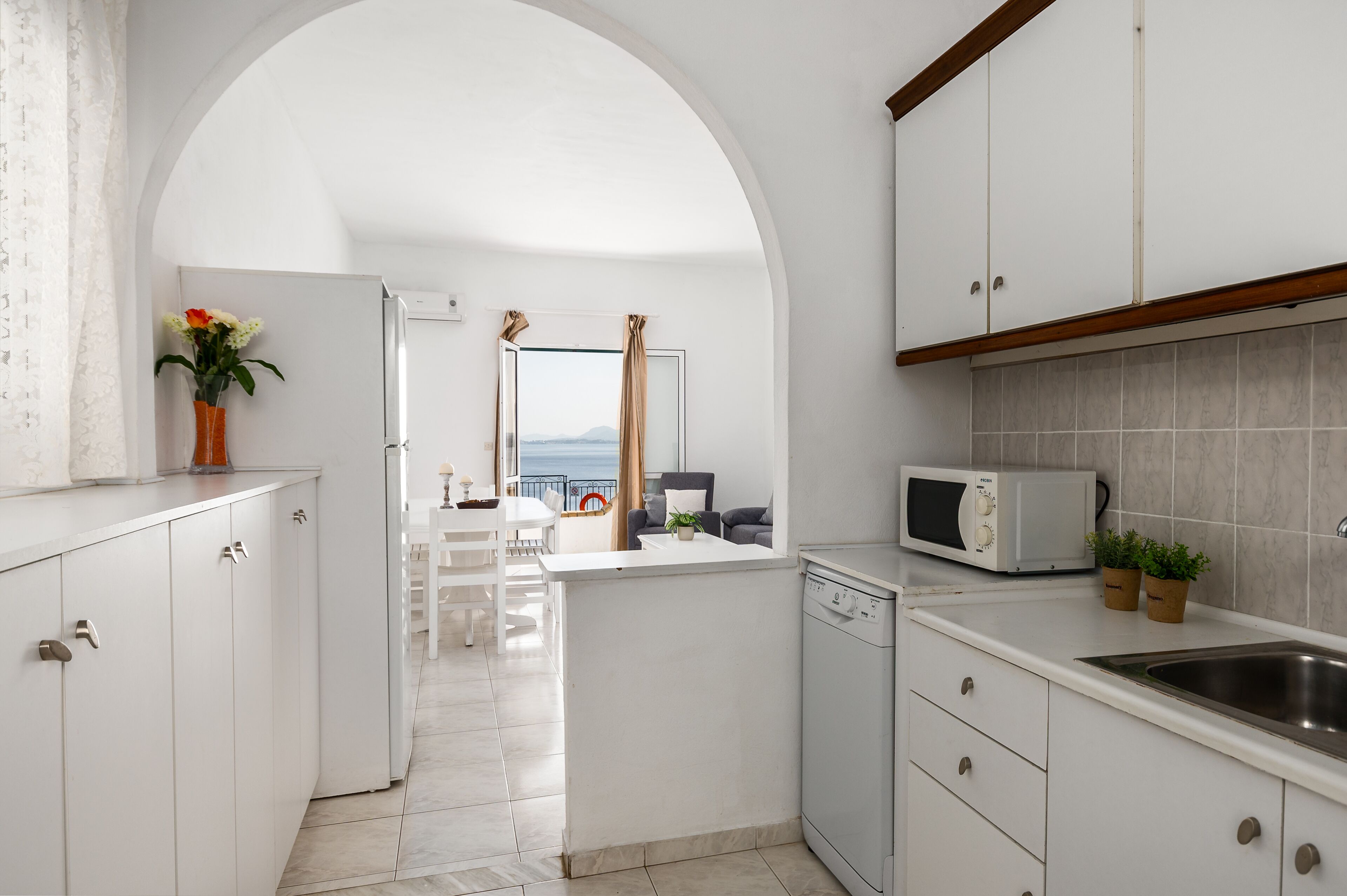 Eine moderne Küche mit weißen Schränken, einem Geschirrspüler, einer Mikrowelle und Blick durch einen Durchgang zu einem Balkon mit Tisch und Stühlen.