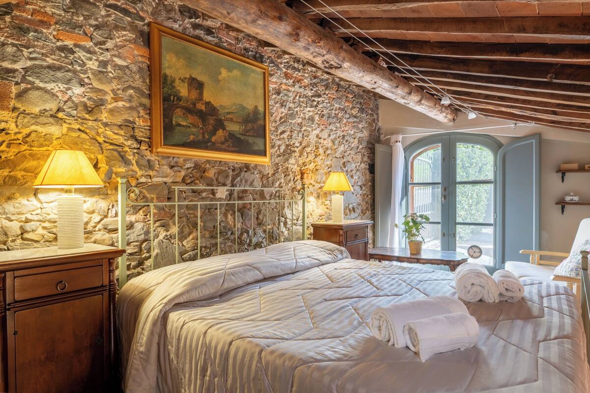 A bedroom with a stone wall, a painting, a bed with white bedding, two bedside lamps, and a window with a view.