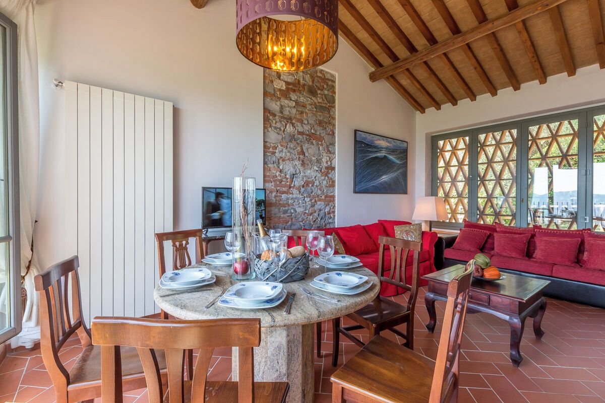 Un coin repas avec une table ronde dressée pour quatre personnes, entourée de chaises en bois. Un pilier en pierre et un canapé rouge sont visibles à l’arrière-plan.