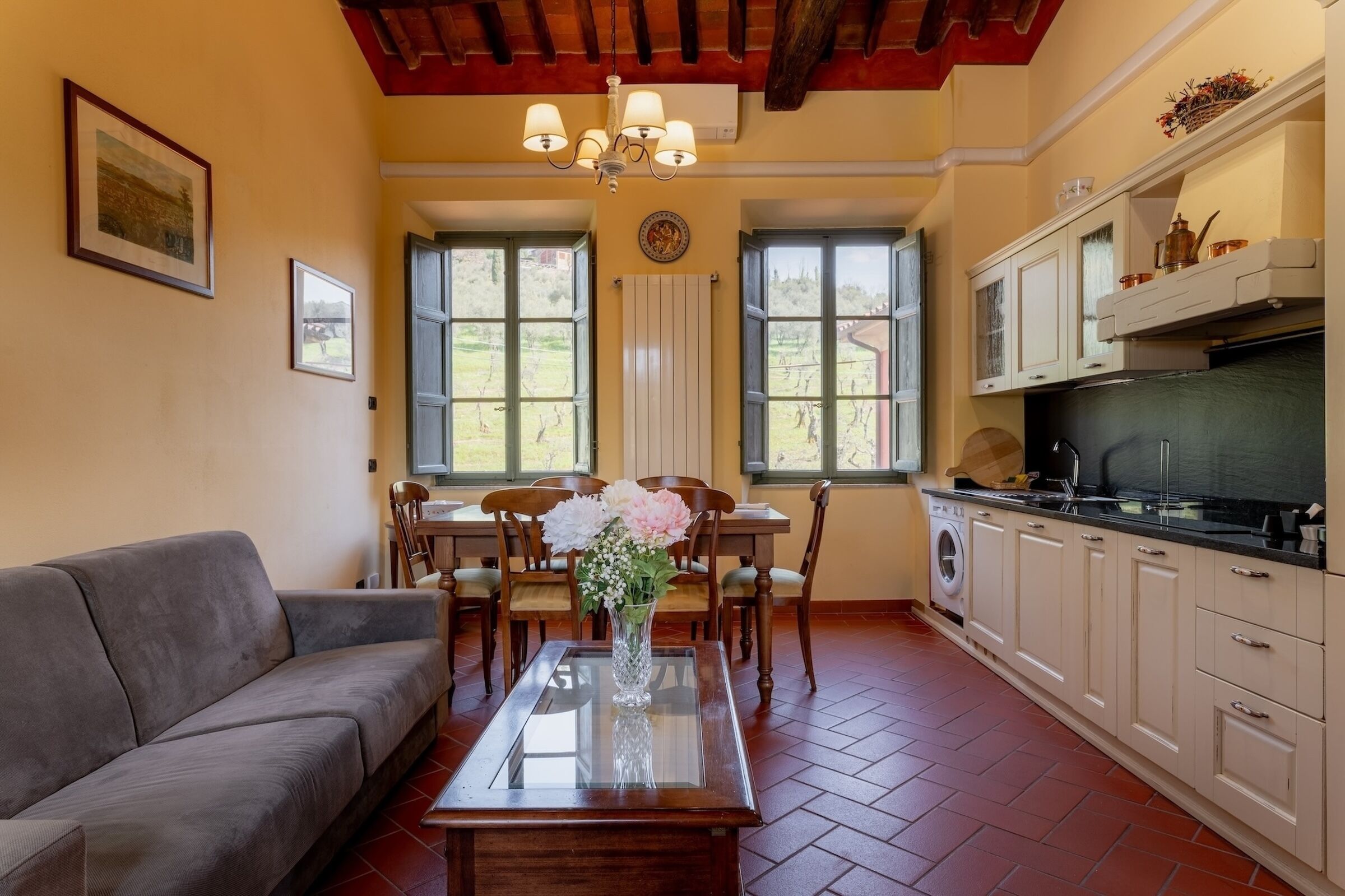 Un soggiorno con divano, tavolo da pranzo, sedie, un vaso con fiori, un lampadario e una cucina con pensili ed elettrodomestici.