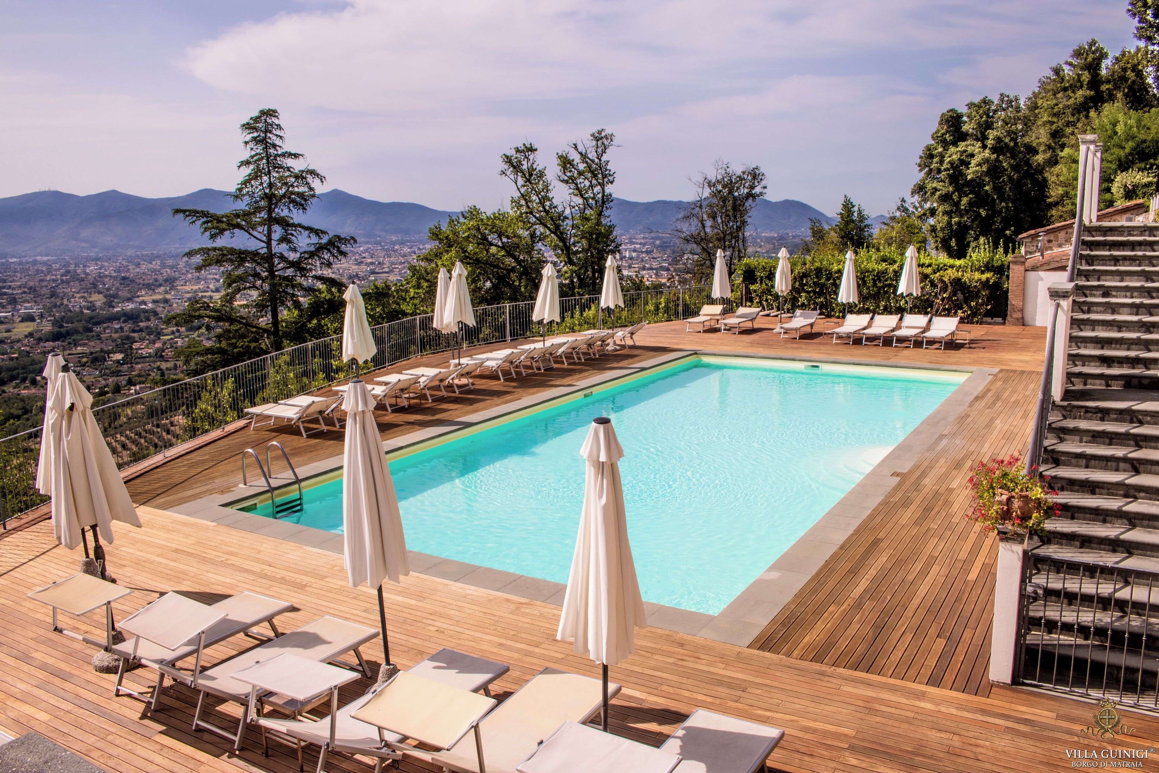 A pool area with lounge chairs and umbrellas, surrounded by a wooden deck and overlooking a cityscape with mountains in the distance.