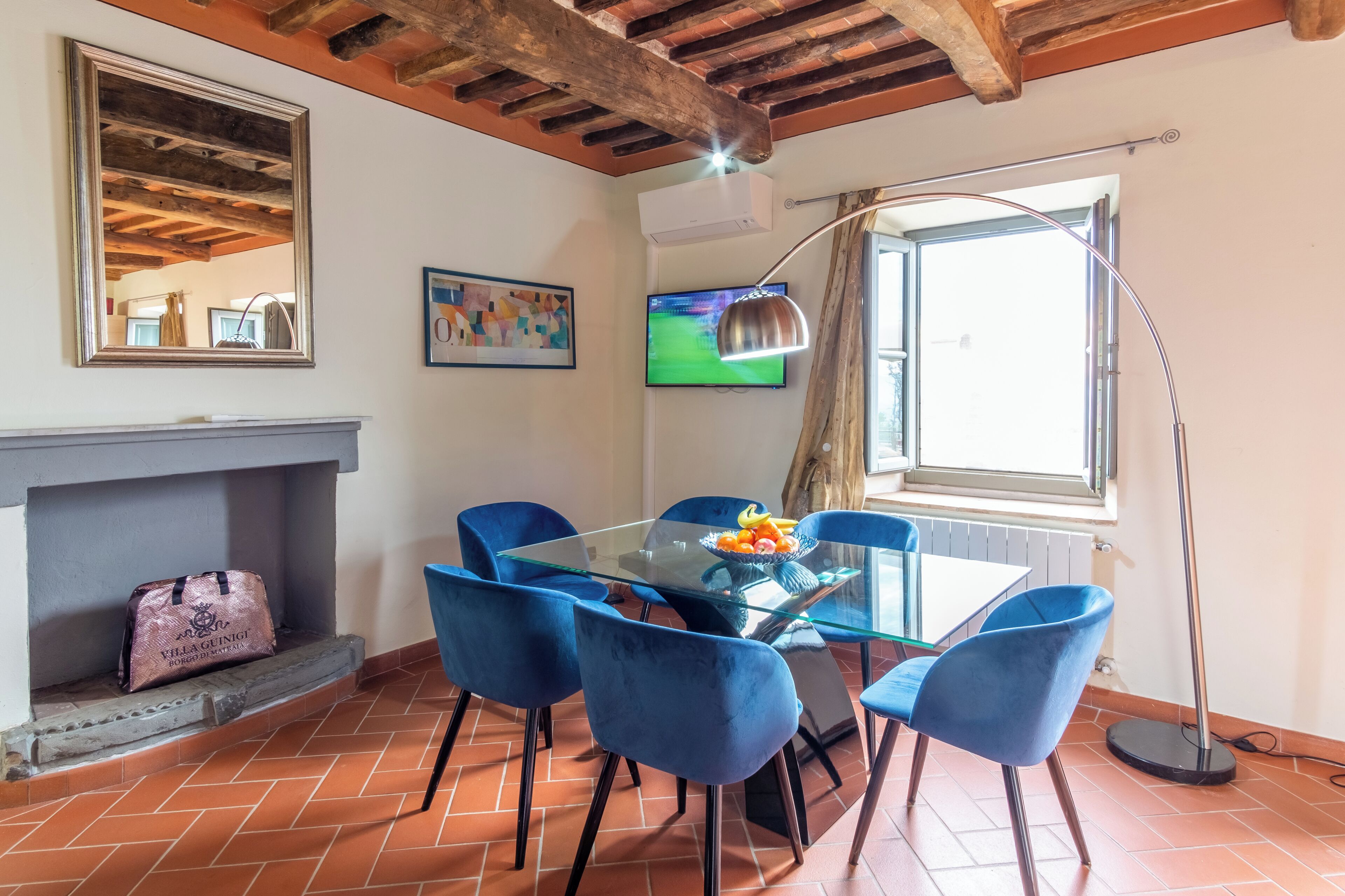 Un área de comedor con mesa de vidrio, sillas azules, chimenea, televisor y un ventanal grande.