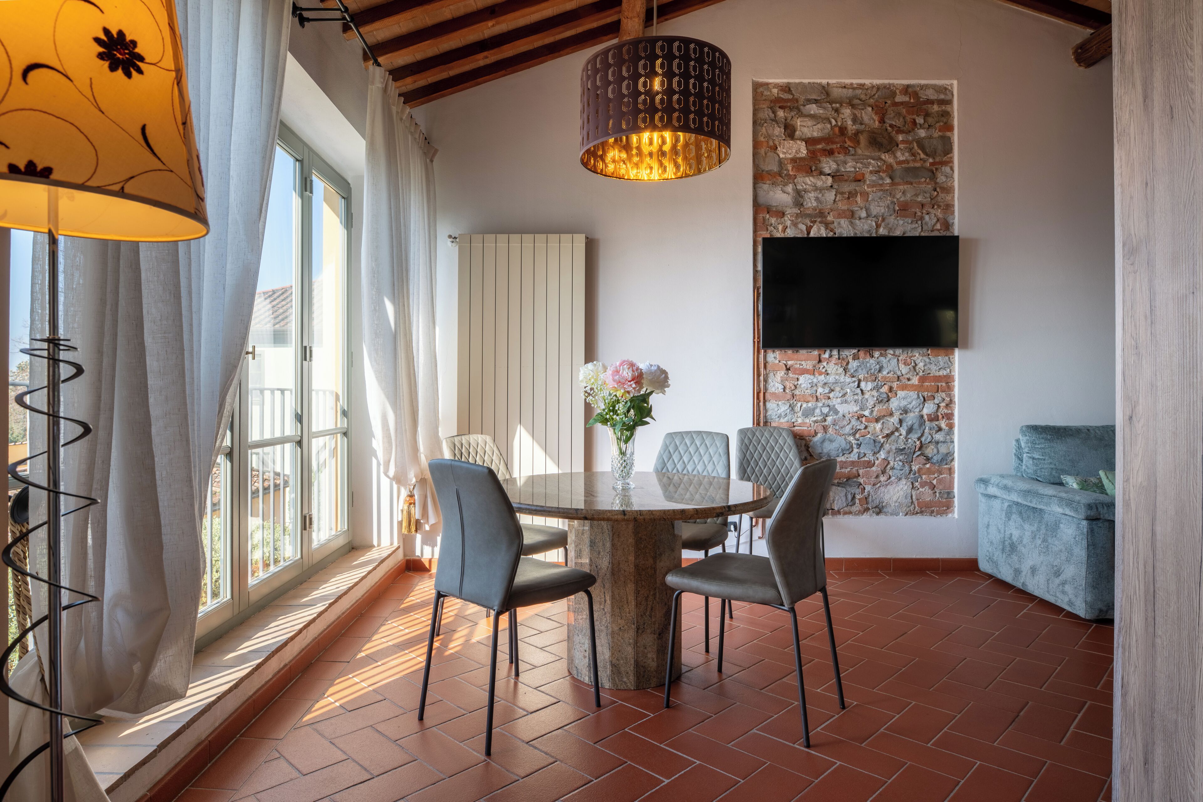A dining area with a round table, four chairs, a vase with flowers, a hanging light fixture, a brick wall, and a television mounted on the wall.