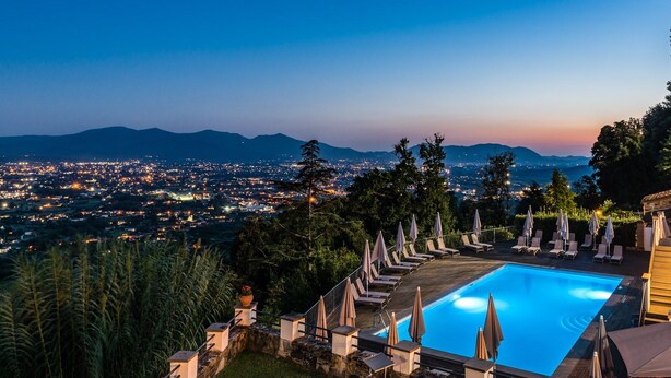 A pool area with lounge chairs and umbrellas, surrounded by greenery and overlooking a cityscape at dusk. - Tenuta Guinigi Antico Borgo di Matraia   (Capannori)