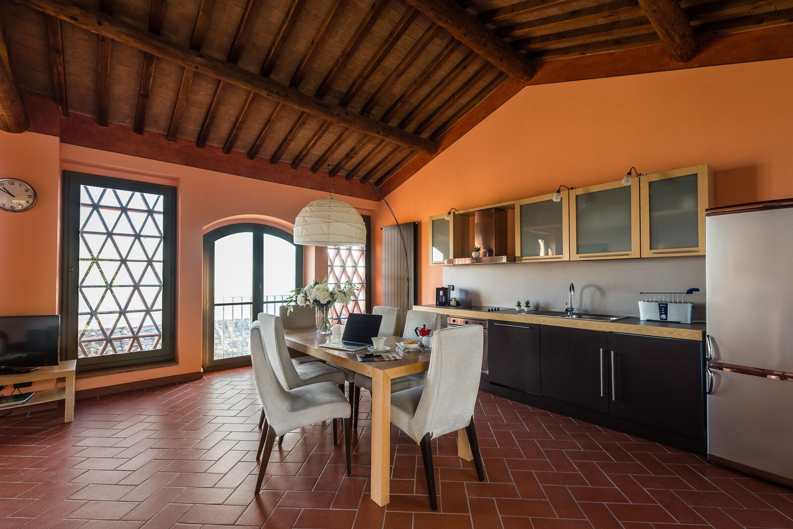 A dining area with a wooden table, chairs, and a laptop. There is a kitchen with cabinets and a refrigerator.