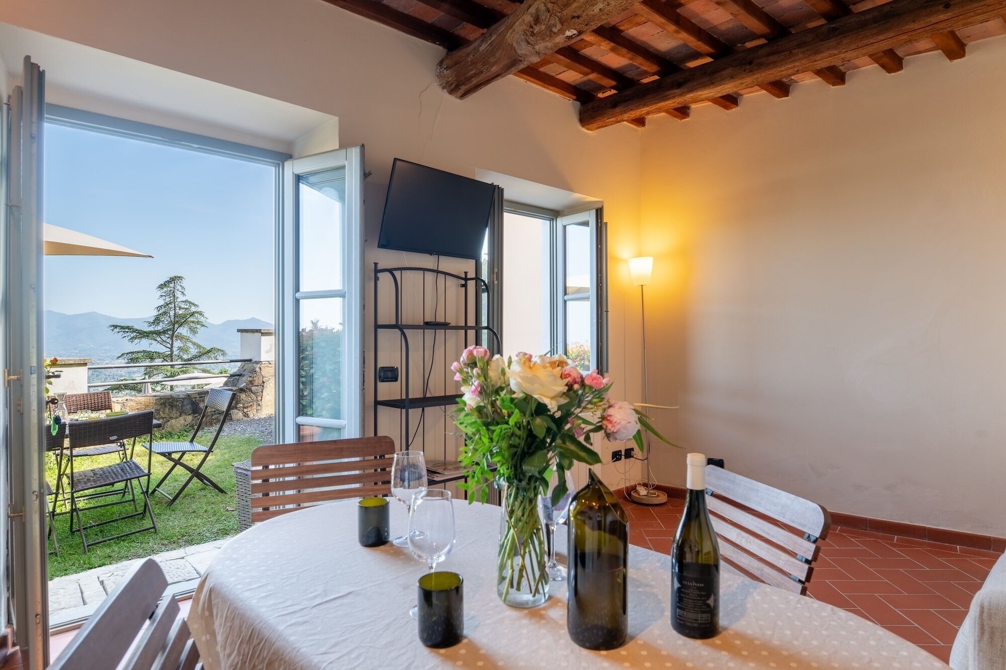 An open terrace with a table set for dining, a vase of flowers, and a bottle of wine.